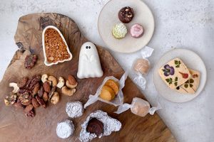 木製プレートに緑茶や紫芋の葛サブレ、桜島小みかんのアマレッティ、花やハーブが飾られた米粉サブレ、ゴースト型の最中スイーツ、ラム酒を使ったベイクドショコラ、ナッツを盛り付けた写真。見た目も可愛らしく、贈り物や配り用にも最適なハロウィンギフト。